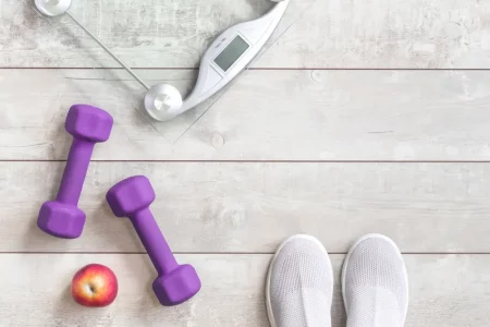 Lightweight modern textile slip on sneakers shoes with weighing scale, trendy purple violet dumbbells and apple on grey wood - Obesity management